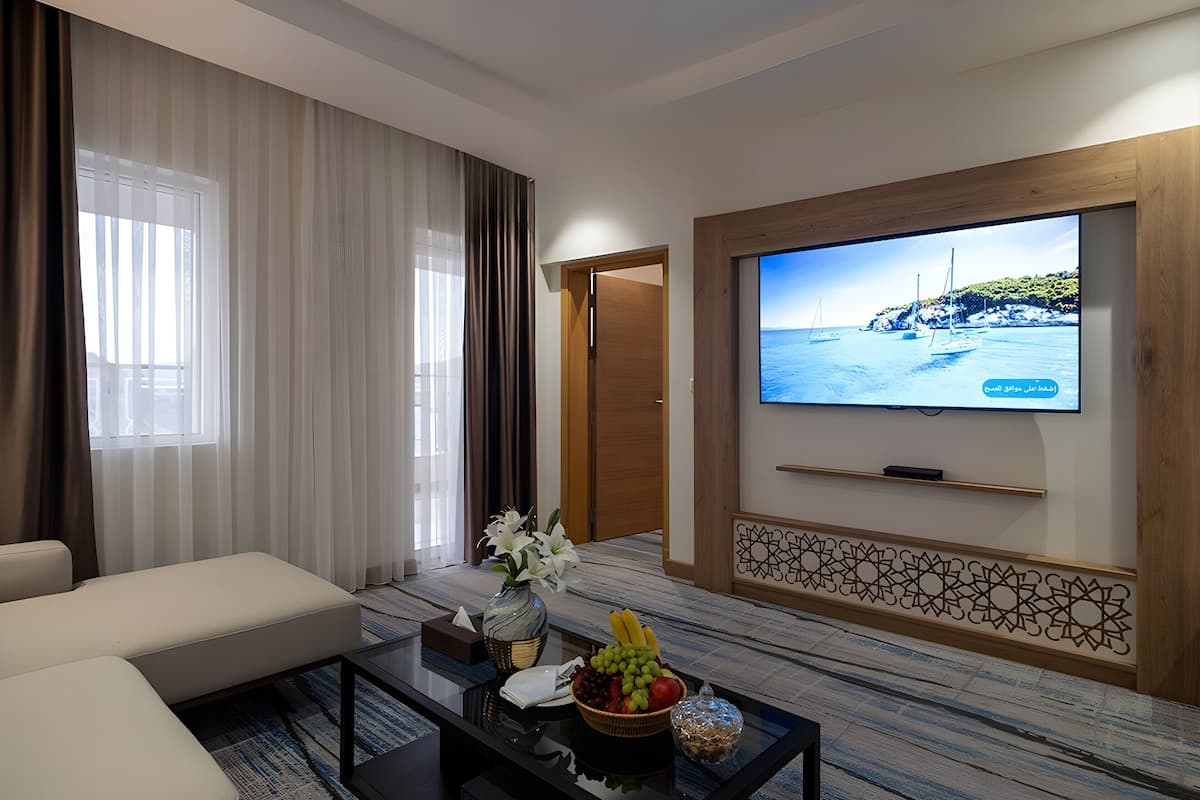 A modern living room with a flat-screen TV mounted on the wall, a comfortable sofa, a coffee table with fruits and a vase, and a patterned carpet.