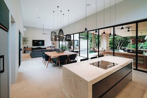 A modern kitchen with a central island, large windows, and a dining area with a sofa and table.