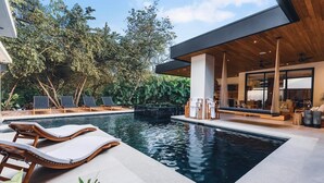 A modern outdoor pool area with lounge chairs, a wooden deck, and a covered seating area with a view of the pool. - Avara House (Reserva de Golf, Hacienda Pinilla)