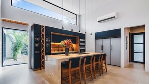 A modern kitchen with a central island, bar stools, and a view of greenery outside. - Avara House (Reserva de Golf, Hacienda Pinilla)