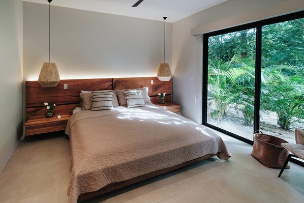 A bedroom with a large bed, two bedside tables, and a view of greenery through a large window.