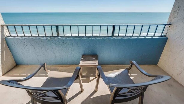 A balcony with two chairs and a small table, overlooking a beach with clear blue water and a metal railing. - Amazing Views, Coastal  Paradise Experience! (Myrtle Beach)