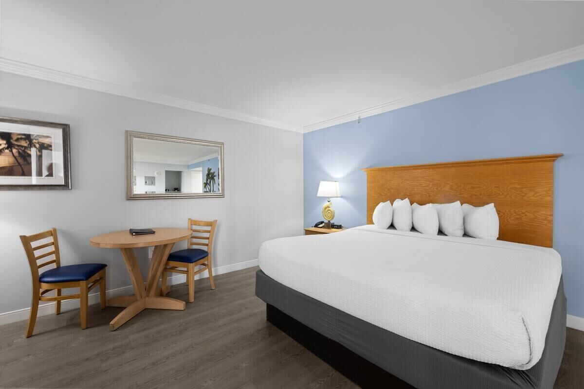 A hotel room with a bed, a round wooden table with two chairs, a lamp, and a mirror on the wall.