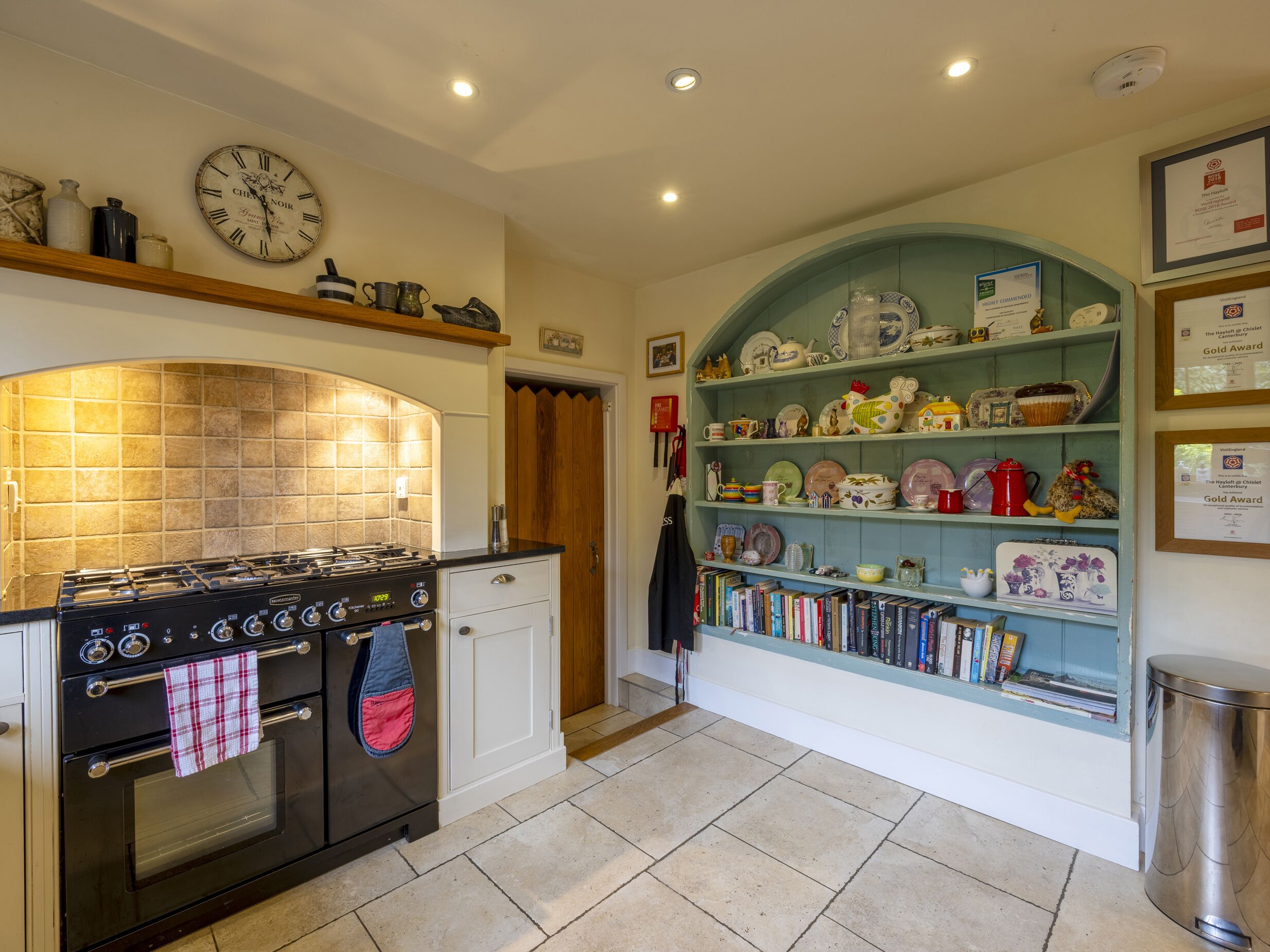 Una cucina con piano cottura, forno e un orologio appeso al muro. Sono presenti scaffali con stoviglie e libri.