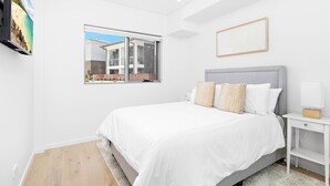 A bedroom with a large bed, a bedside table, a nightstand, a window with a view of a building, and a framed picture on the wall. - Wine Down - Small family getaway (Kiama)