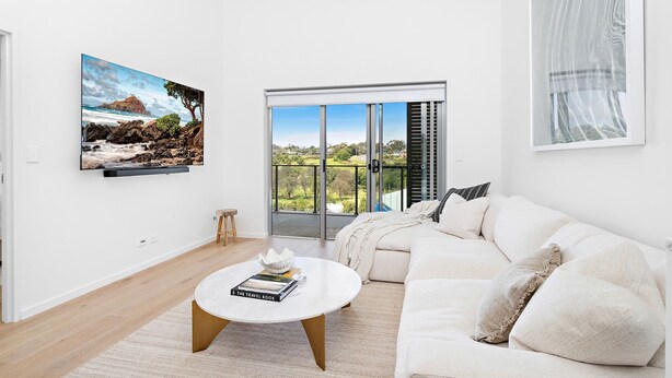A modern living room with a white sofa, a round coffee table with books, a painting of a coastal landscape, and a large sliding glass door leading to a balcony with a view of greenery. - Wine Down - Small family getaway (Kiama)