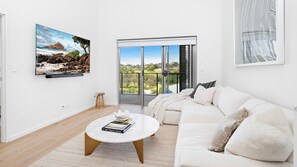 A modern living room with a white sofa, a round coffee table with books, a painting of a coastal landscape, and a large sliding glass door leading to a balcony with a view of greenery. - Wine Down - Small family getaway (Kiama)