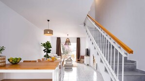 Ein modernes Wohnzimmer mit einem Holzesstisch, weißen Regalen, einer Treppe mit weißen Geländern und einem Blick ins Freie durch große Fenster.