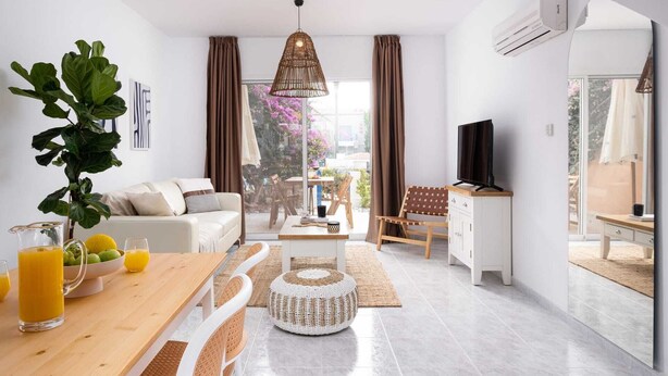 Ein modernes Wohnzimmer mit Sofa, Esstisch und Fernseher. Ein großes Fenster mit Vorhängen, eine Pendelleuchte und eine Pflanze sind ebenfalls zu sehen.