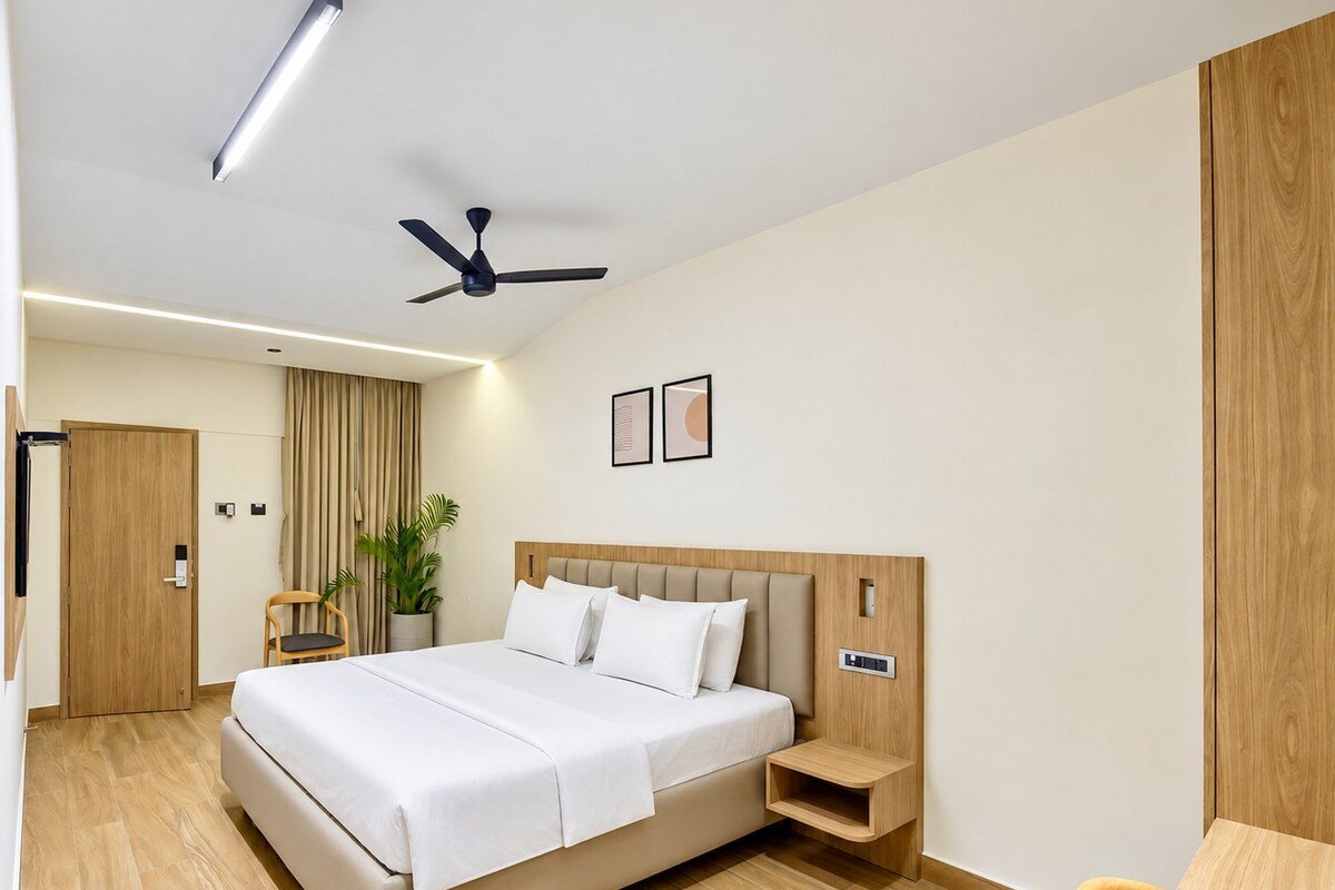 A hotel room with a bed, bedside table, chair, ceiling fan, and a potted plant.
