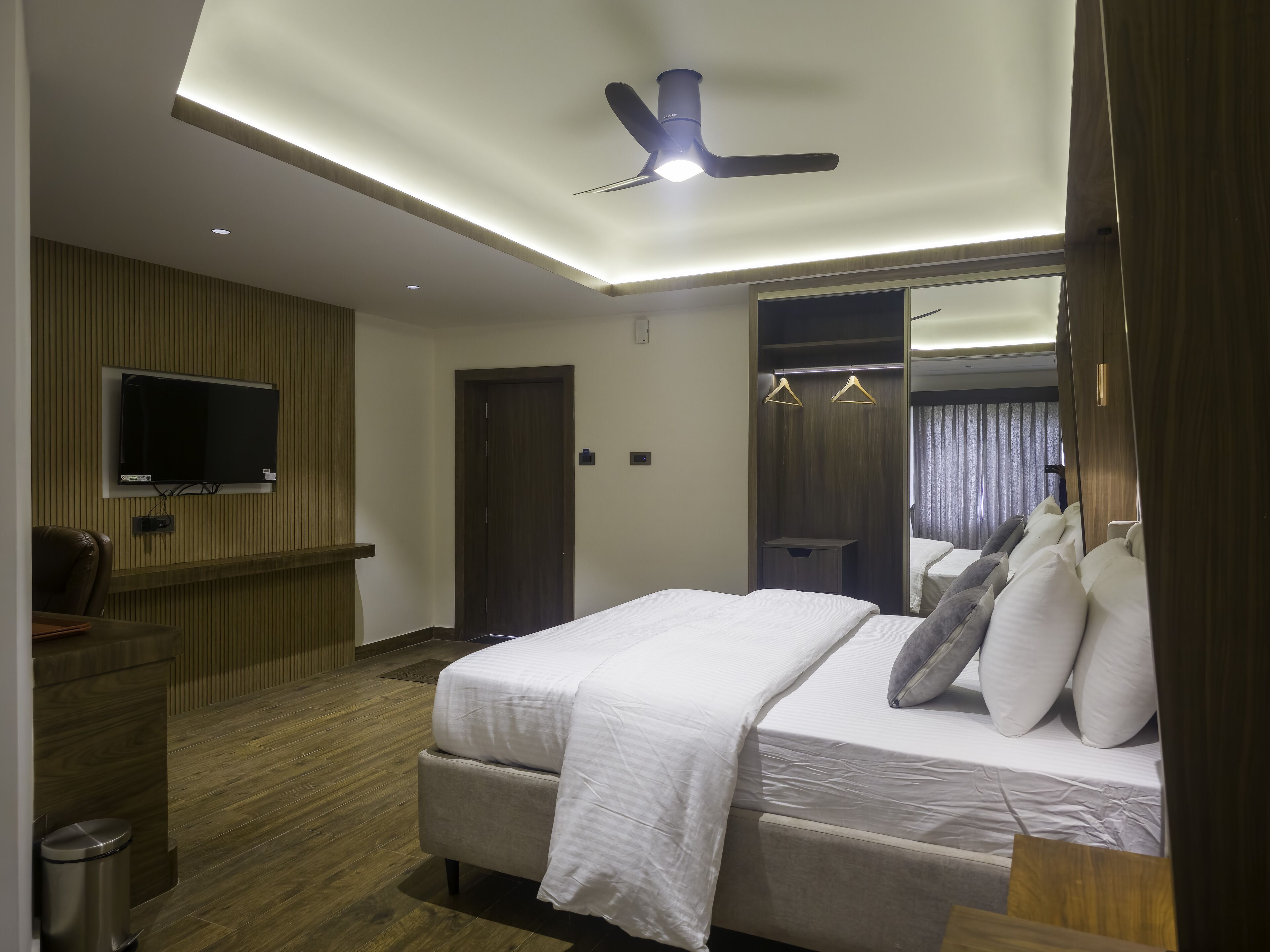 A modern hotel room with a large bed, a TV mounted on the wall, a ceiling fan, and a wardrobe with a mirror.