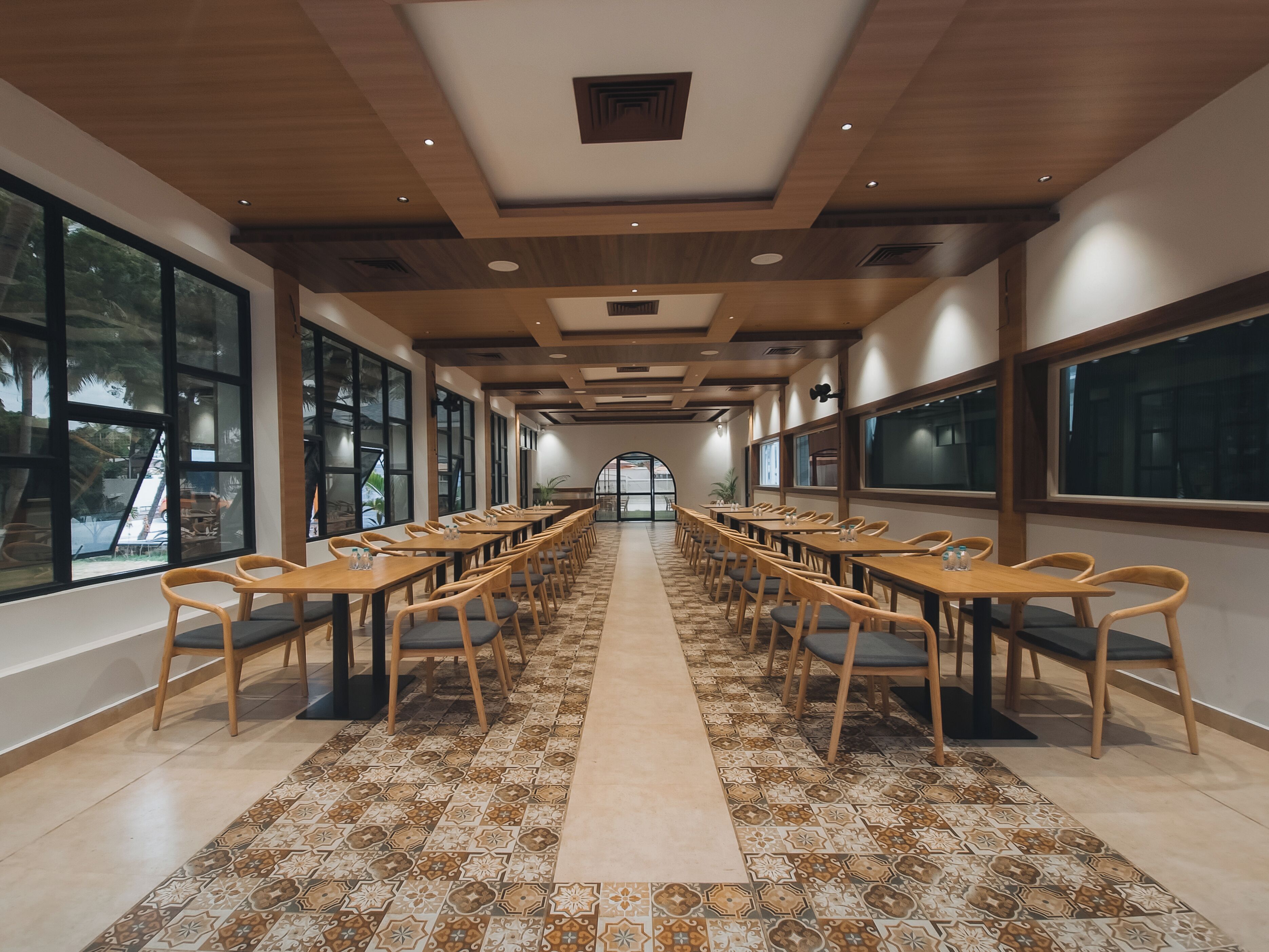 A long dining hall with wooden tables and chairs, large windows, and a patterned floor.