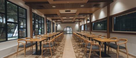 A long dining hall with wooden tables and chairs, large windows, and a patterned floor.