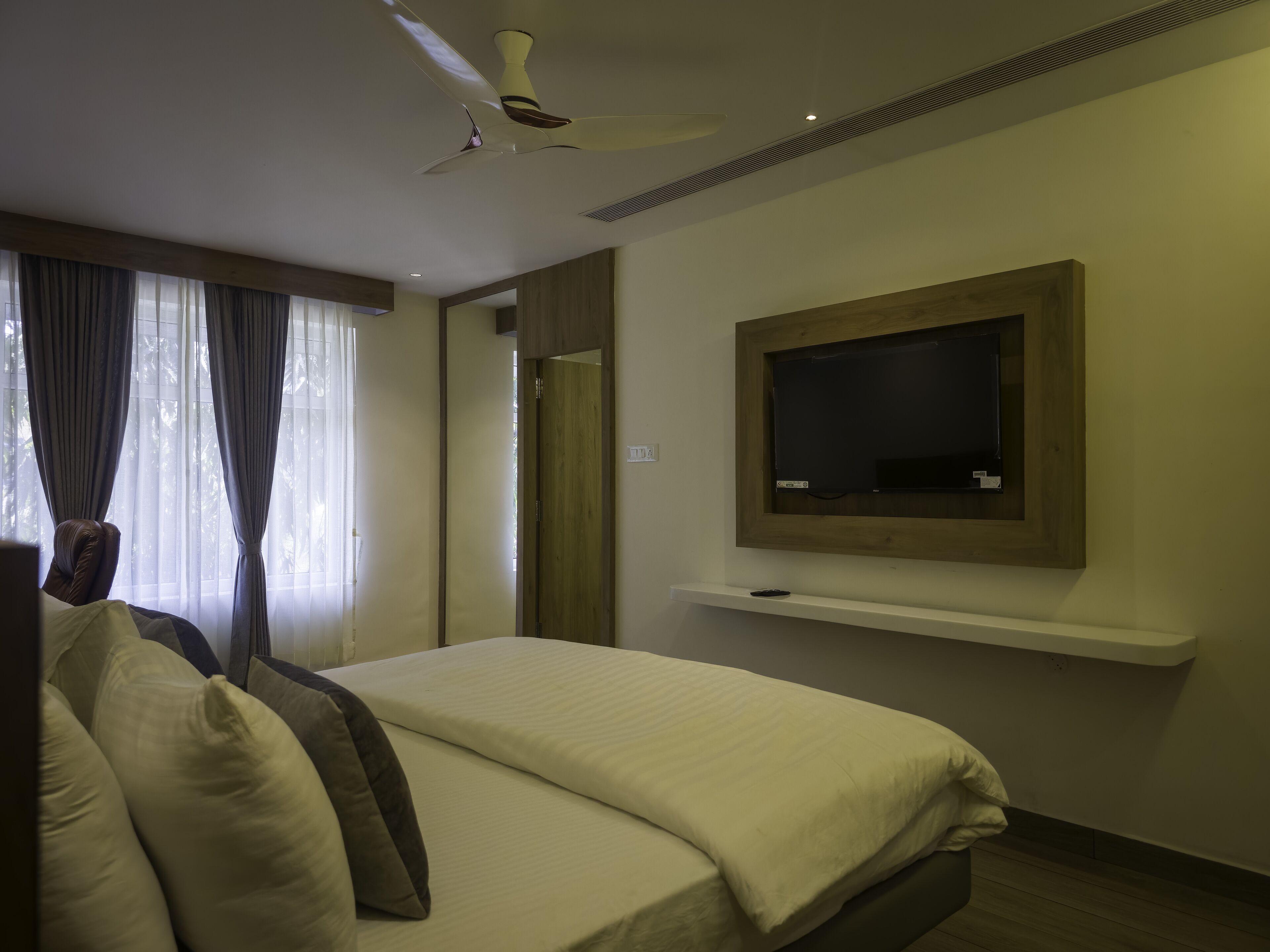 Une chambre d’hôtel avec un lit, un ventilateur de plafond, un téléviseur fixé au mur et une fenêtre avec des rideaux.