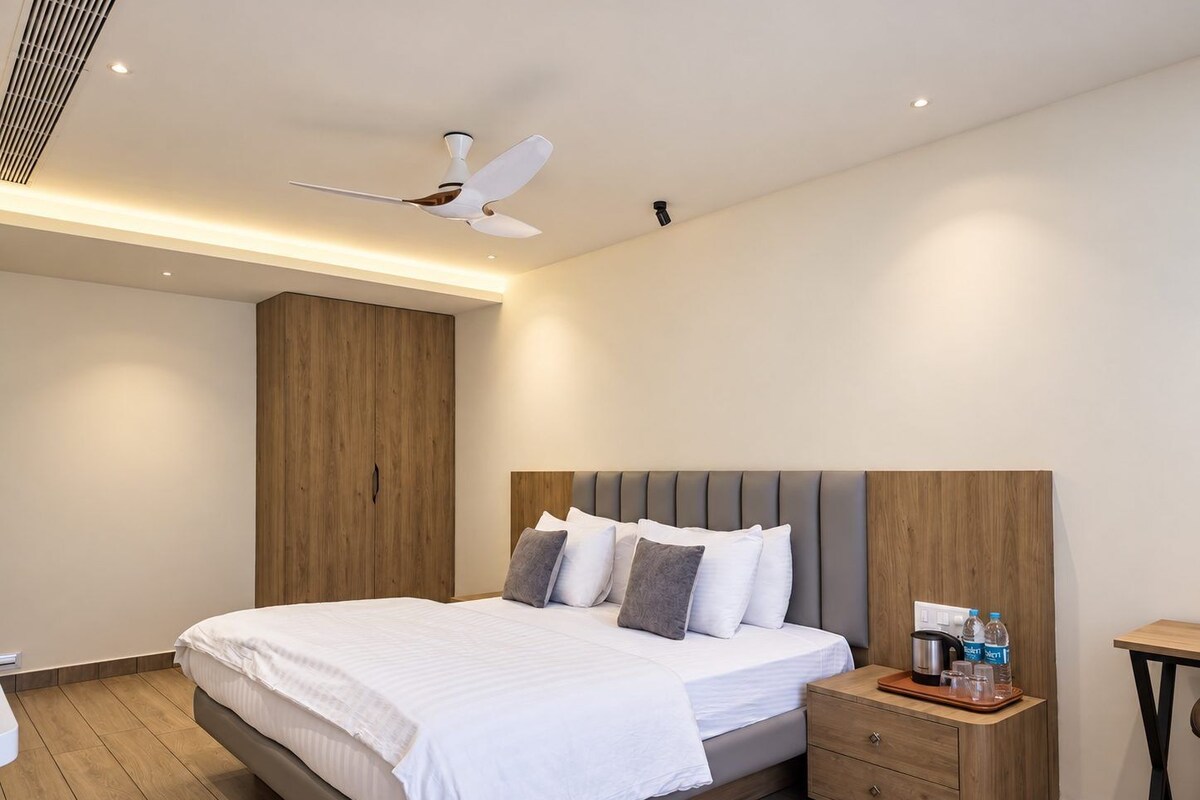 A modern hotel room with a large bed, a wooden wardrobe, a bedside table with a coffee maker, and a ceiling fan.