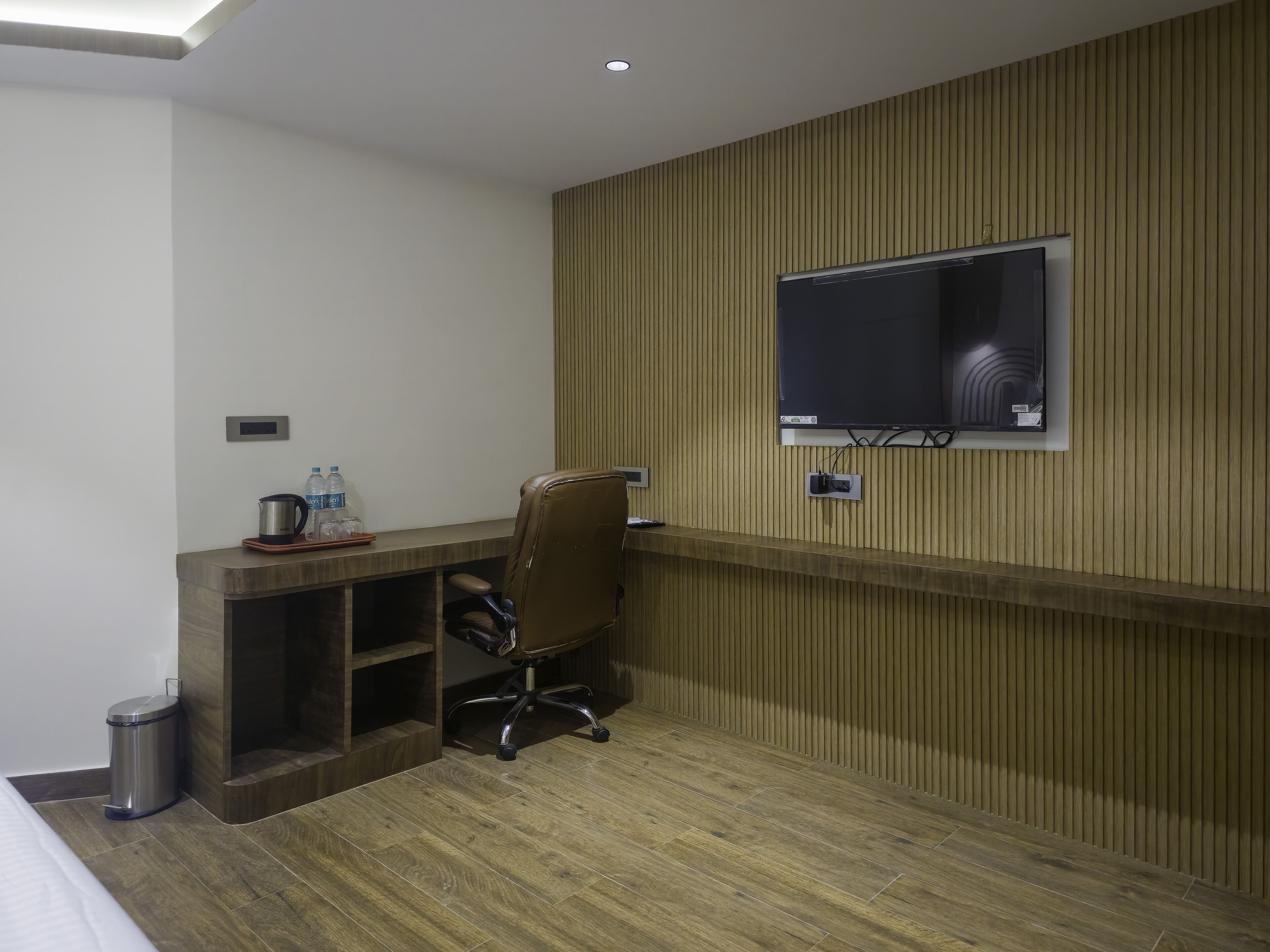 A hotel room with a wooden panel wall, a mounted TV, a desk with a chair, a trash bin, and a bed.