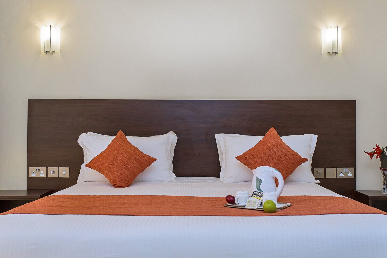Una cama bien hecha con un cabecero marrón, dos almohadas naranjas y una bandeja con una taza y fruta en la mesita de noche.