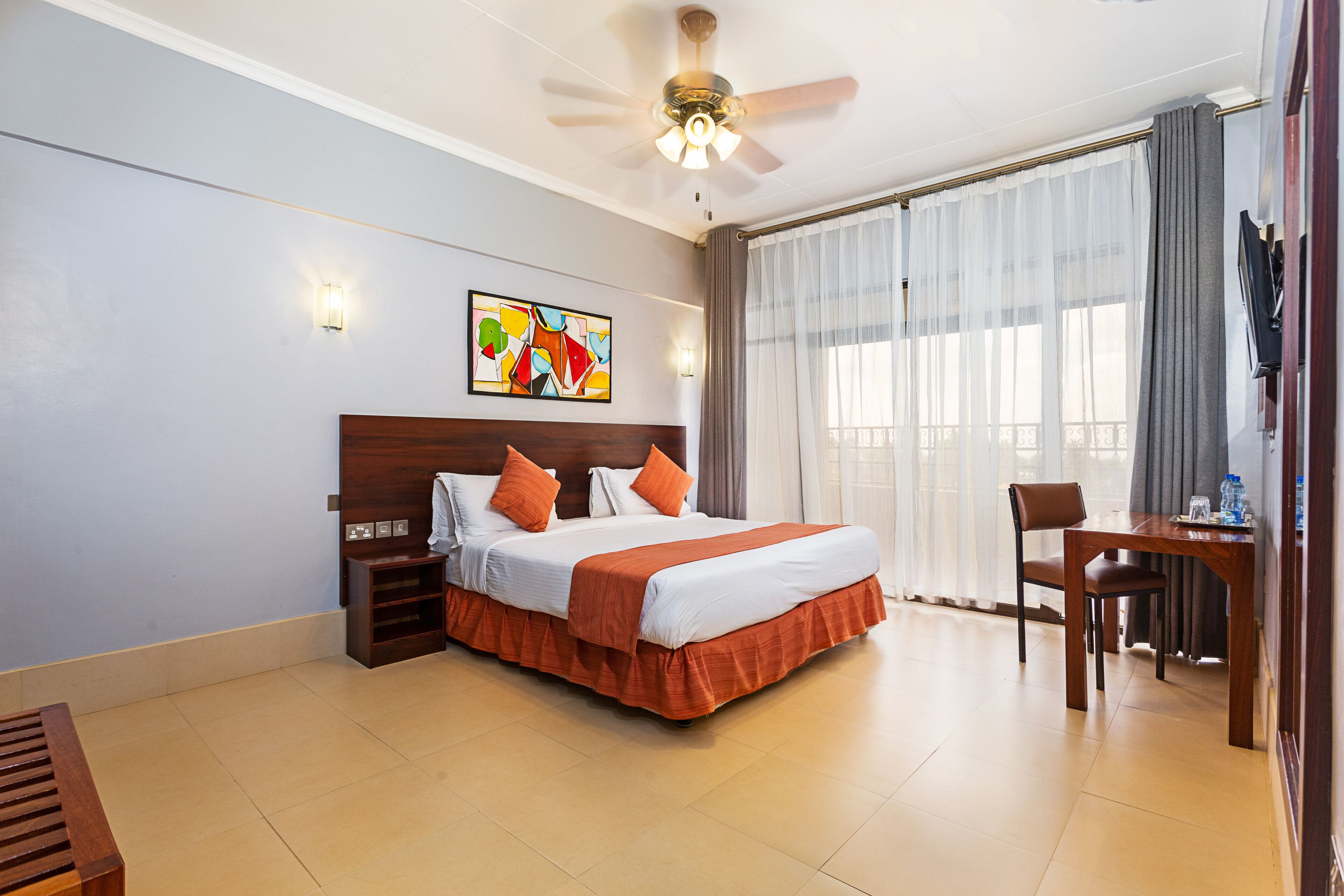 A hotel room with a large bed, a desk, a chair, a ceiling fan, and a window with curtains.