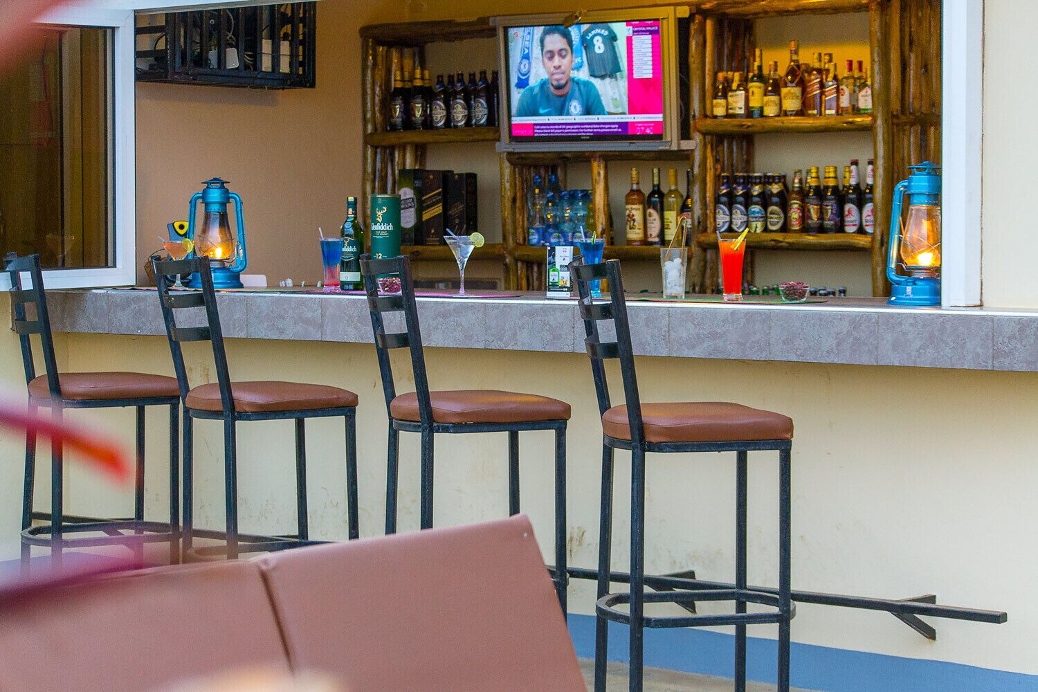 A bar area with four stools, a television screen, and shelves stocked with bottles.