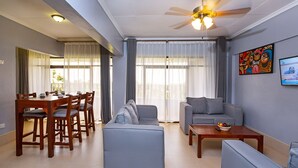 A spacious living room with a dining area, grey sofas, a ceiling fan, and large windows with sheer curtains. - Roswam Hotel (Kerugoya)