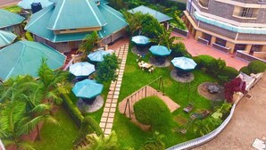 A courtyard with multiple green-roofed buildings, outdoor seating areas, and a central pathway. - Roswam Hotel (Kerugoya)
