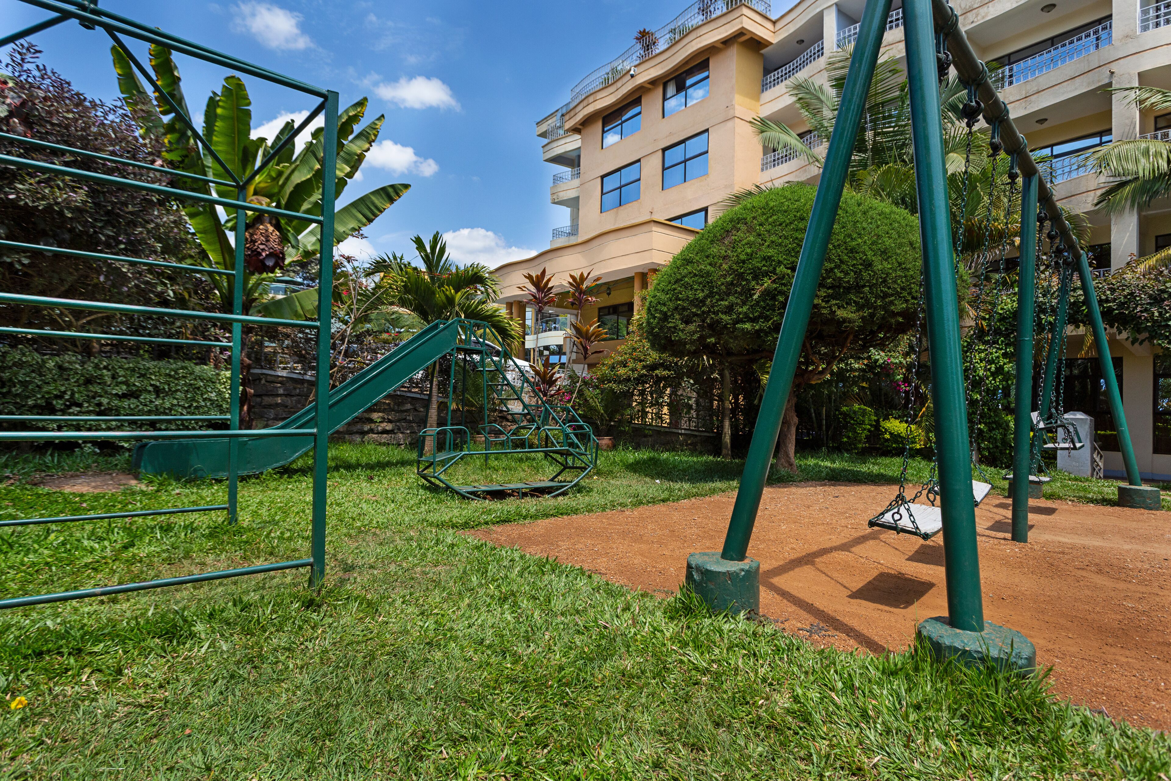 A playground with swings and a slide, surrounded by greenery and a building in the background.