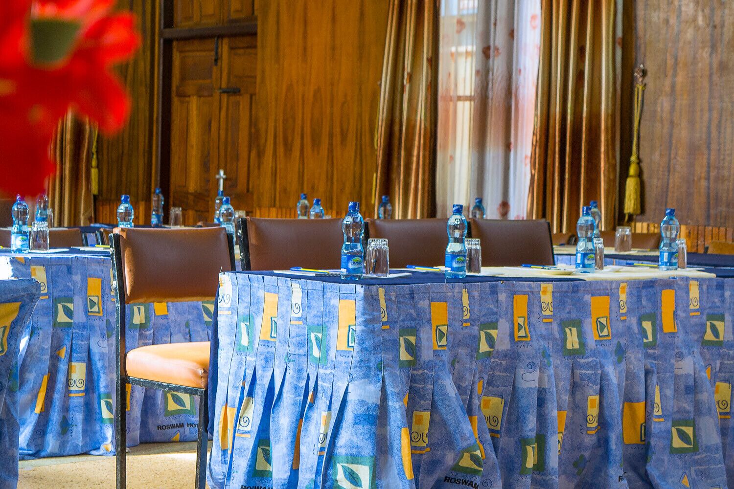 Un salón de conferencias con mesas cubiertas con manteles estampados, sillas y botellas de agua en cada una.