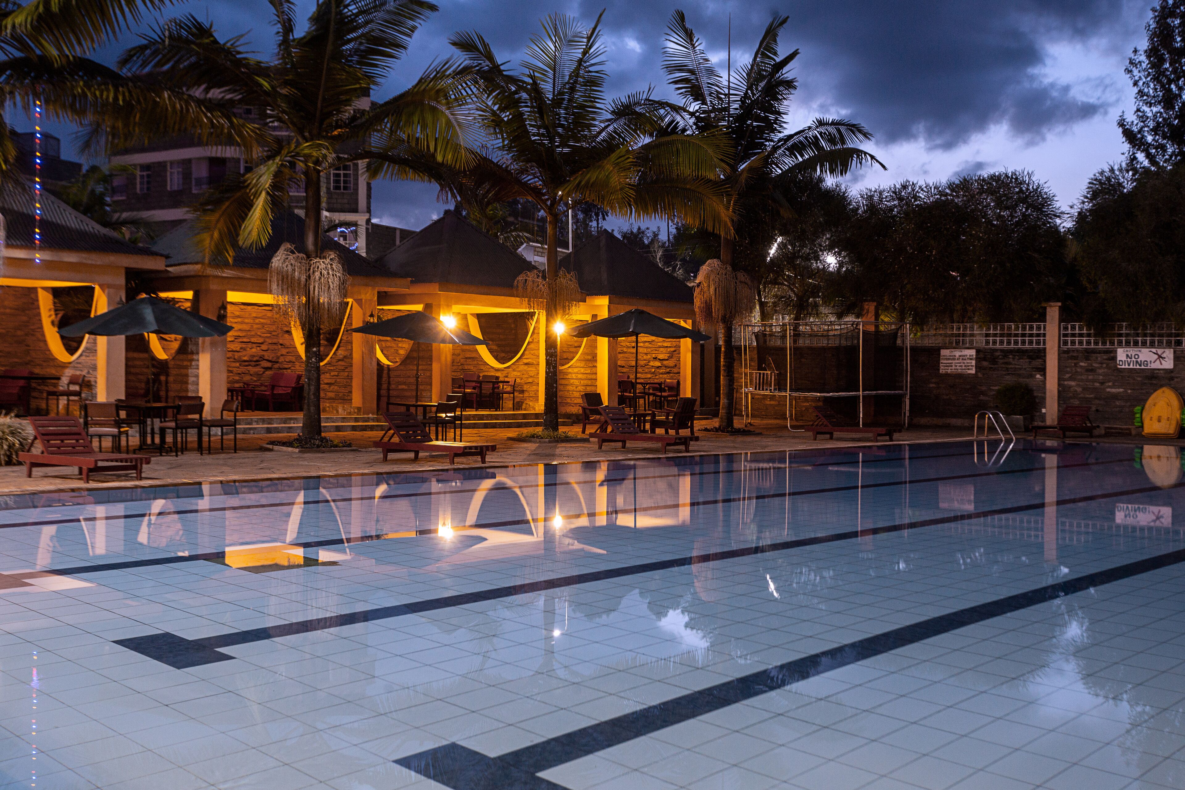A swimming pool with lounge chairs and umbrellas, surrounded by palm trees and a building with stone walls and warm lighting.