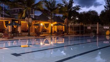 A swimming pool with lounge chairs and umbrellas, surrounded by palm trees and a building with stone walls and warm lighting.