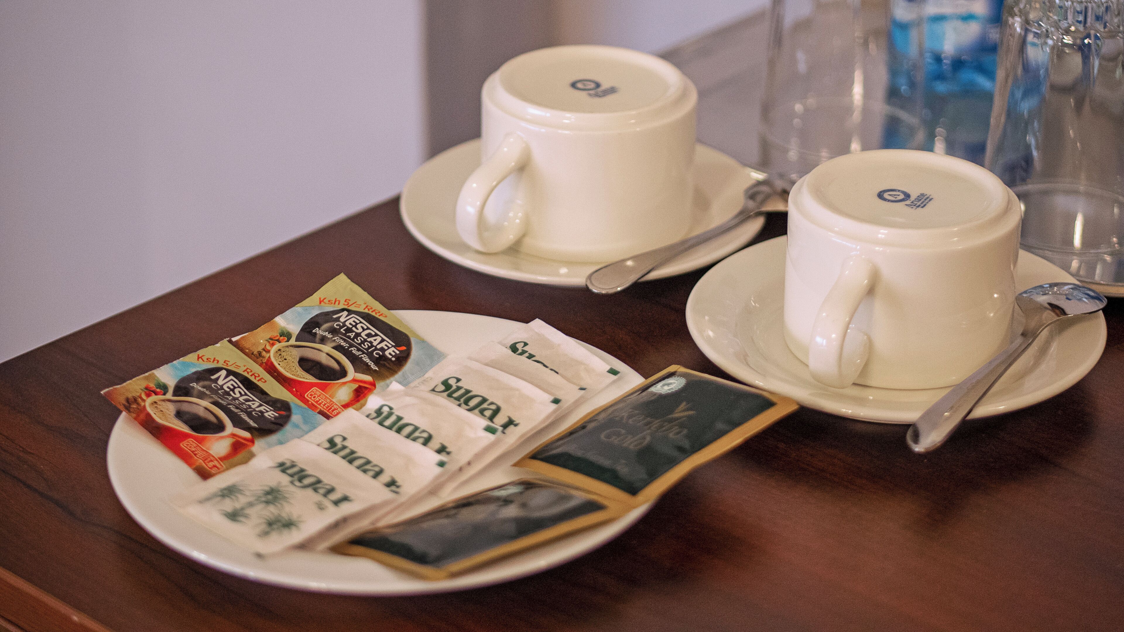 Una mesa con dos tazas y platillos blancos, un plato con sobres de café y un recipiente de vidrio.