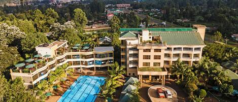 A multi-story hotel with a swimming pool, surrounded by lush greenery and a view of the cityscape.