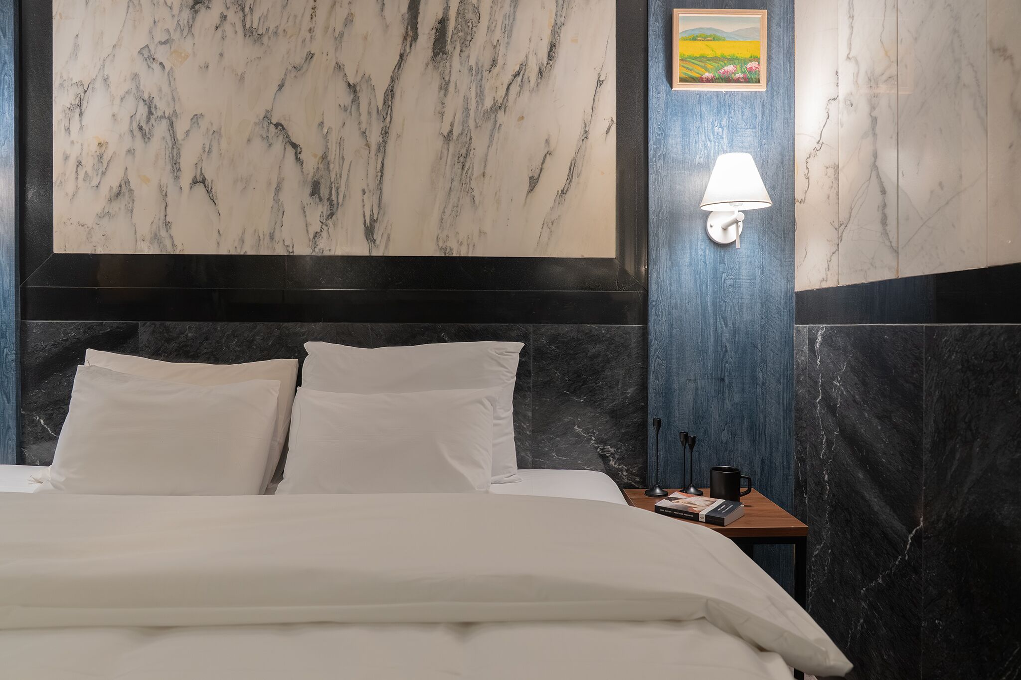 A hotel room with a bed, white pillows, a nightstand with a lamp, and a picture on the wall.