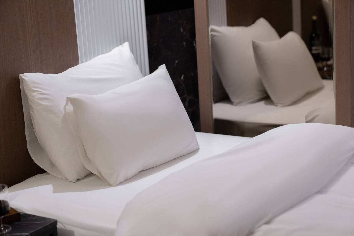 A neatly made bed with white linens and pillows, a wooden headboard, and a mirror reflecting part of the room.