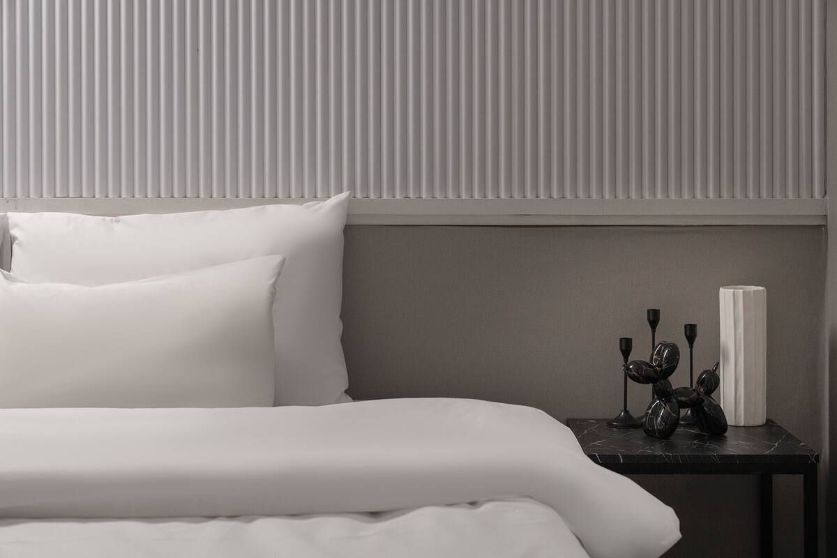 A neatly made bed with white linens, a dark wooden nightstand with a vase and decorative items, and a wall with vertical blinds.