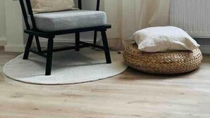 A room with a wooden floor, a black chair with a cushion, a woven basket with a cushion, and a window with sheer curtains. - Schöner Wohnen im Stadtkern (Paderborn)