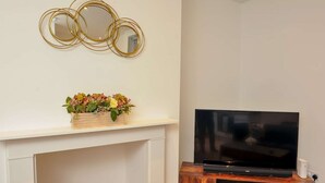 A living room with a fireplace, a flat-screen TV on a wooden stand, and a floral arrangement on the mantel.