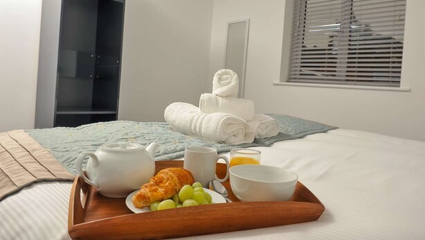 Un lit bien fait, avec un plateau contenant divers éléments pour le petit-déjeuner, notamment des croissants, des raisins et du jus, ainsi qu’une théière blanche.