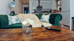 Un salon avec un canapé vert, une table basse en bois, un vase avec des fleurs blanches et une télécommande.