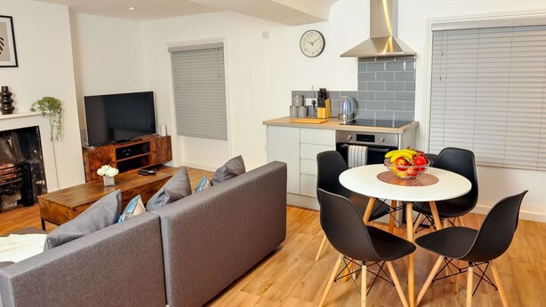 A modern living room with a grey sofa, a TV, and a fireplace. There is a kitchen area with a stove, sink, and a fruit bowl on the table. - 50 Queens Road Apartment 3 (Reading)