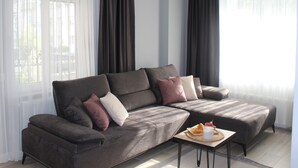 A modern living room with a sectional sofa, a small side table, and a window with sheer curtains. - Cosy Suite Hotel (Konyaalti)