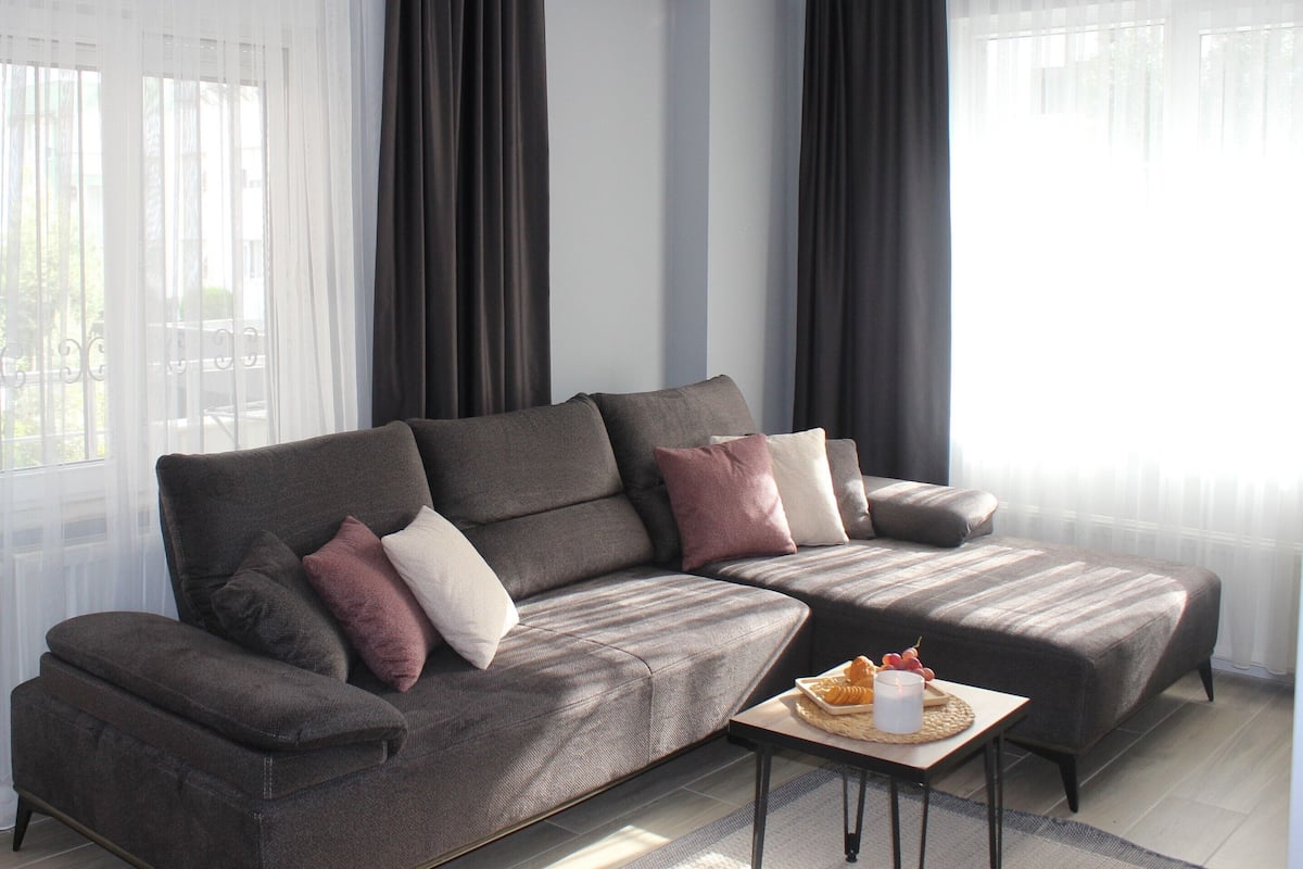 A modern living room with a sectional sofa, a small side table, and a window with sheer curtains.