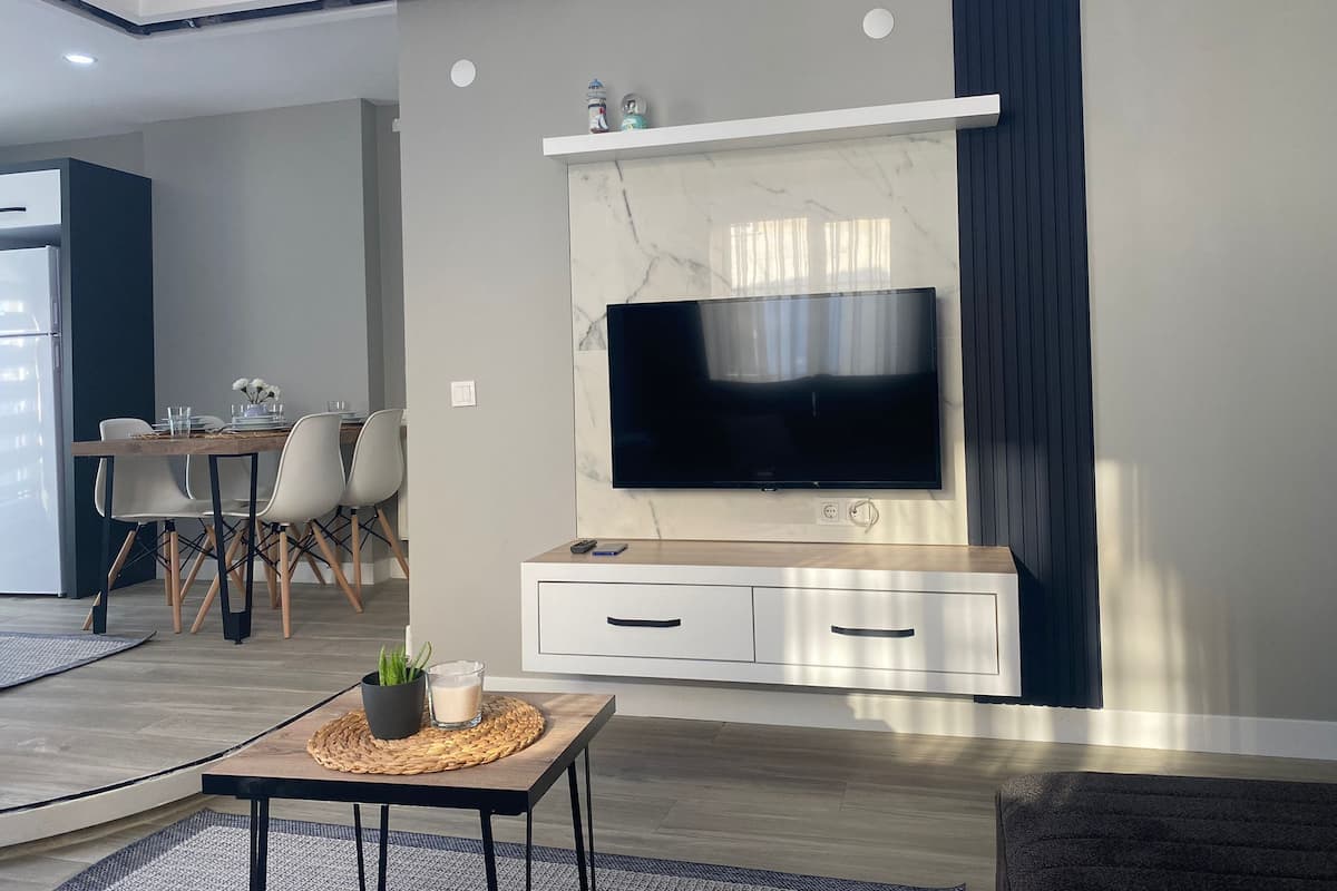 A modern living room with a large flat-screen TV mounted on a marble wall, a dining area with a table and chairs, and a small side table with a plant and candle.