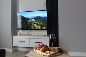 Una sala de estar moderna con un televisor de pantalla plana mostrando un paisaje pintoresco, una mesa de madera con aperitivos, una copa de vino y un control remoto.