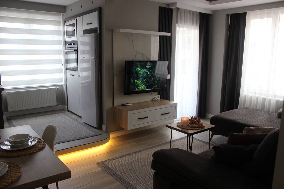 A modern living room with a flat-screen TV, a sofa, a dining table with a candle, and a kitchen area with a microwave and oven.