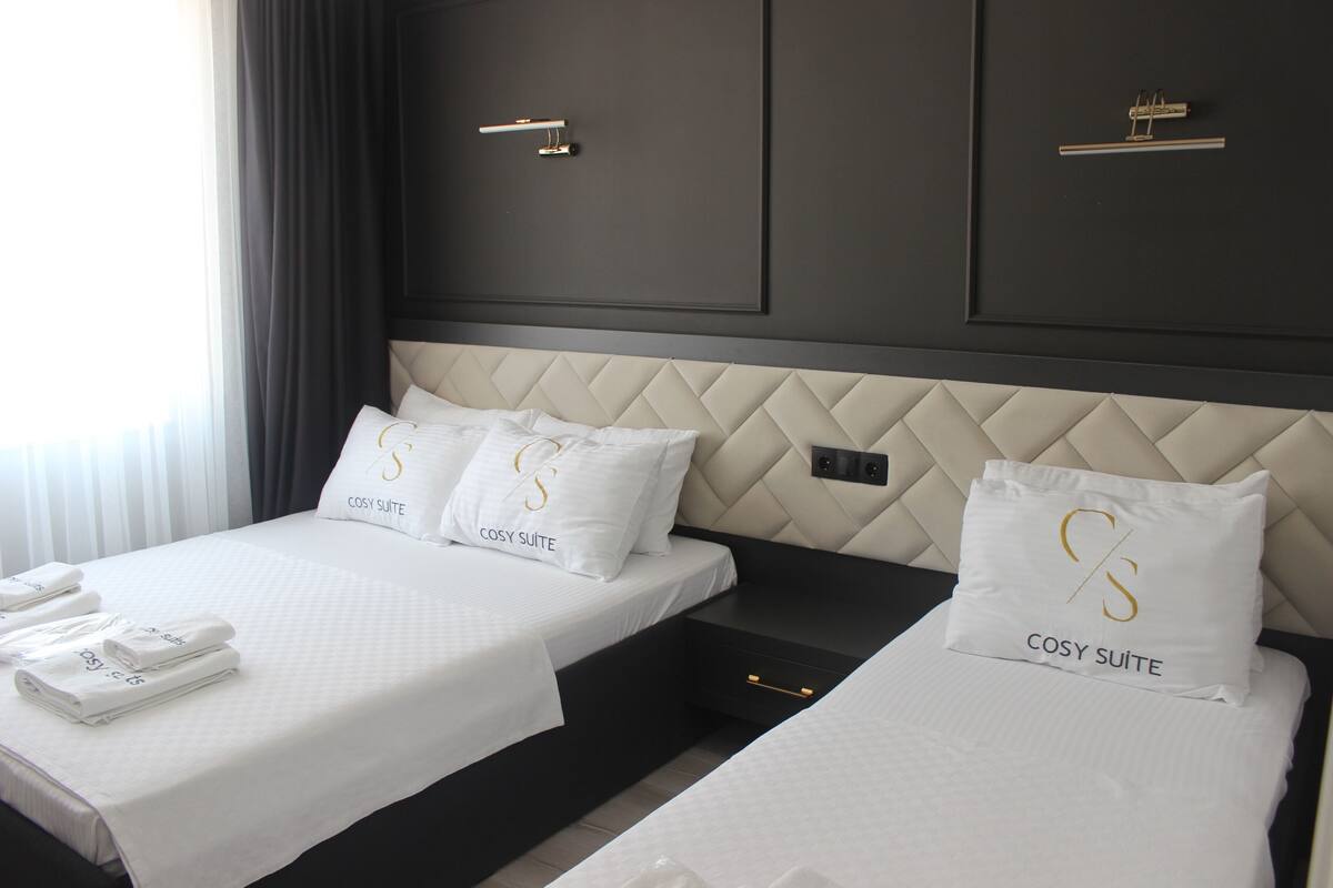 A hotel room with two beds, each with a headboard and pillows labeled "COSY SUITE". There are folded towels on the beds and a small drawer under the beds.