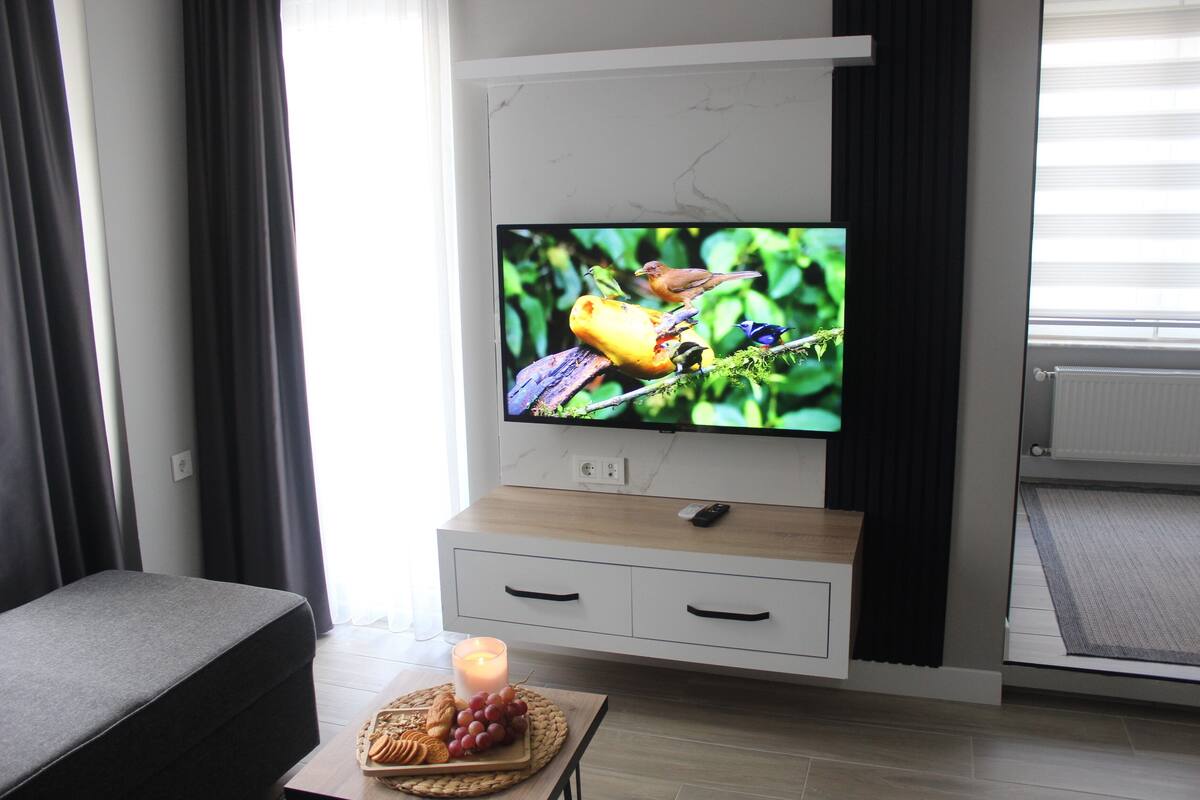 A modern living room with a flat-screen TV mounted on a white cabinet, a grey sofa, a small table with snacks, and a lit candle.