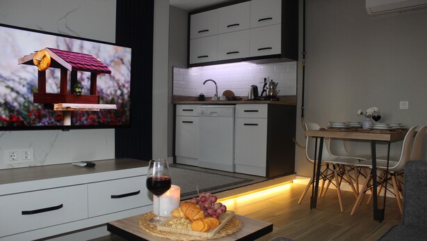 A modern kitchen with white cabinets, a dining area with a table set for two, and a television displaying a birdhouse. - Cosy Suite Hotel (Konyaalti)