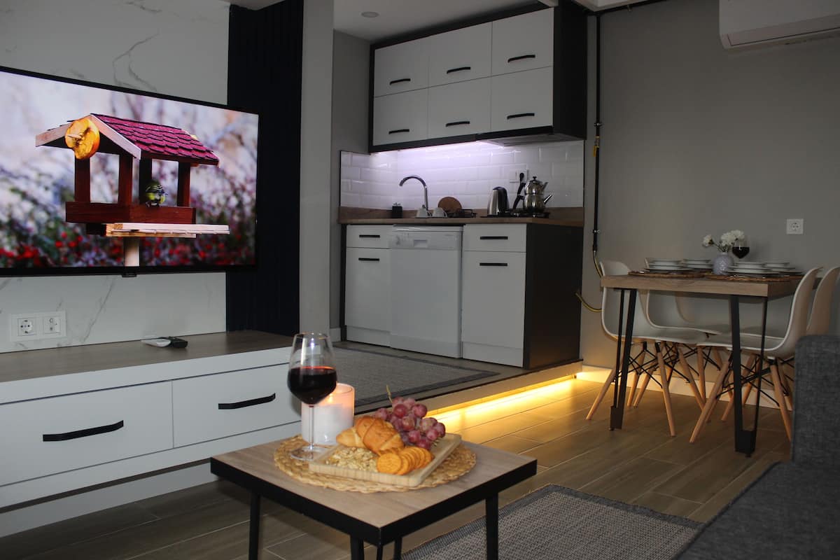 A modern kitchen with white cabinets, a dining area with a table set for two, and a television displaying a birdhouse.
