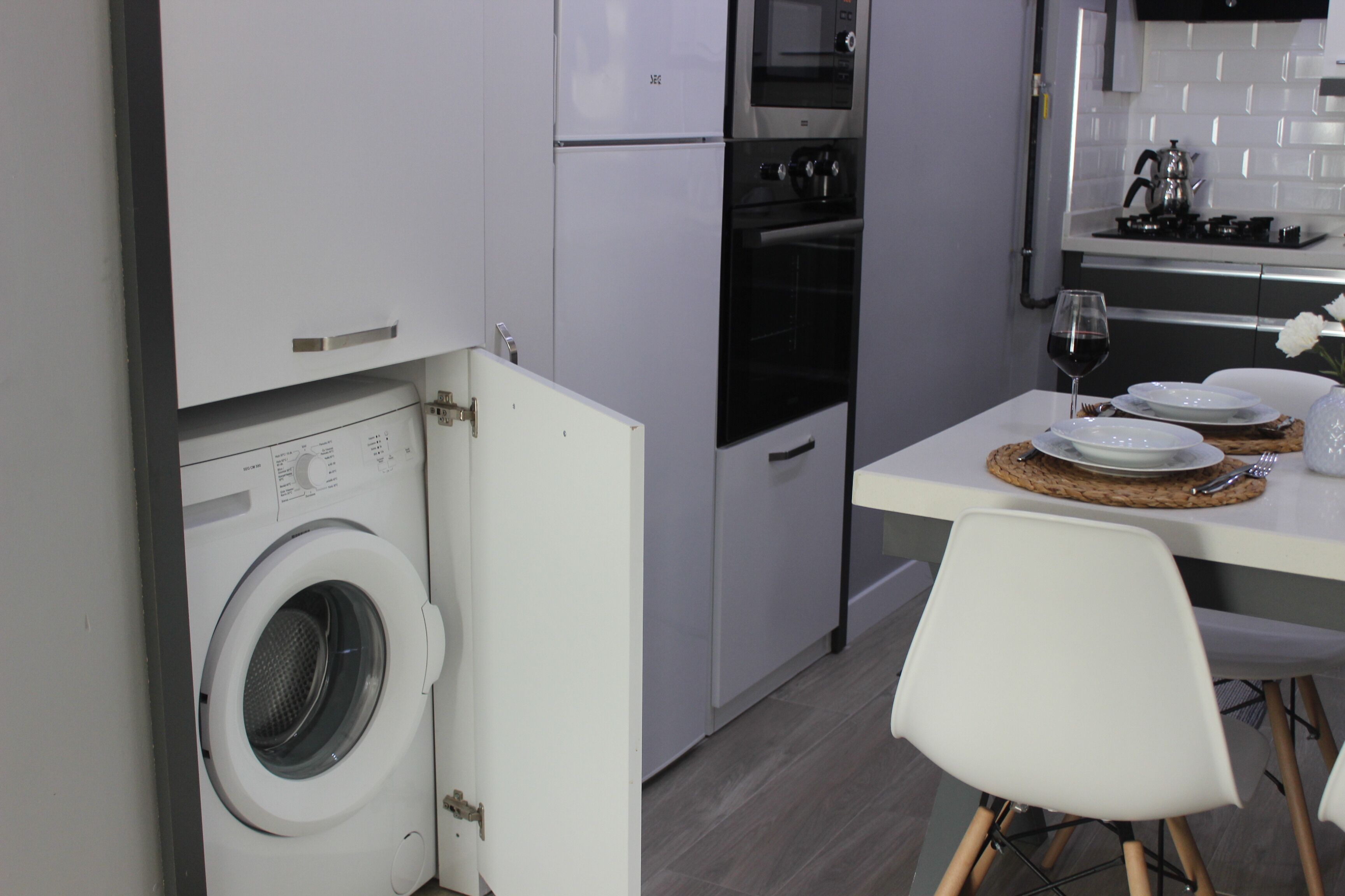 A compact kitchen with a built-in washer, a dining area with a table set for two, and a white chair.