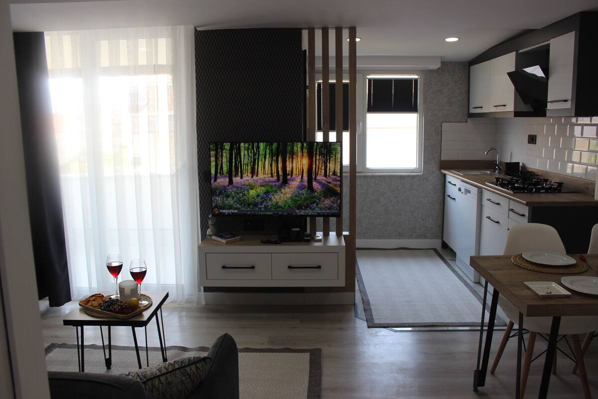A modern living room with a flat-screen TV, a dining area with a table set for two, and a kitchen with white cabinets and a stove.